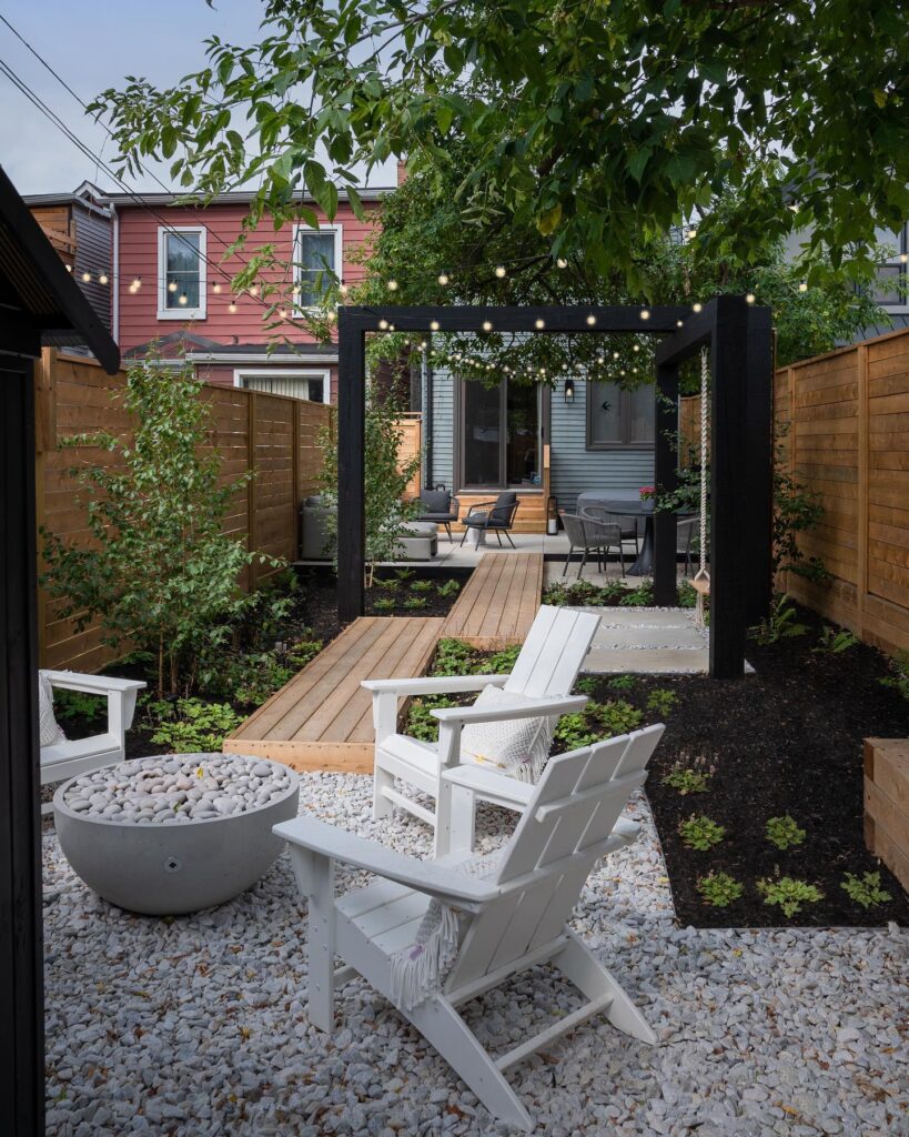 Cozy backyard corner with modern gray seating, a round table, and black chairs, leading to a secluded hammock and fire pit area. The space is surrounded by lush greenery, wooden fencing, and soft string lighting, creating an intimate, inviting retreat.